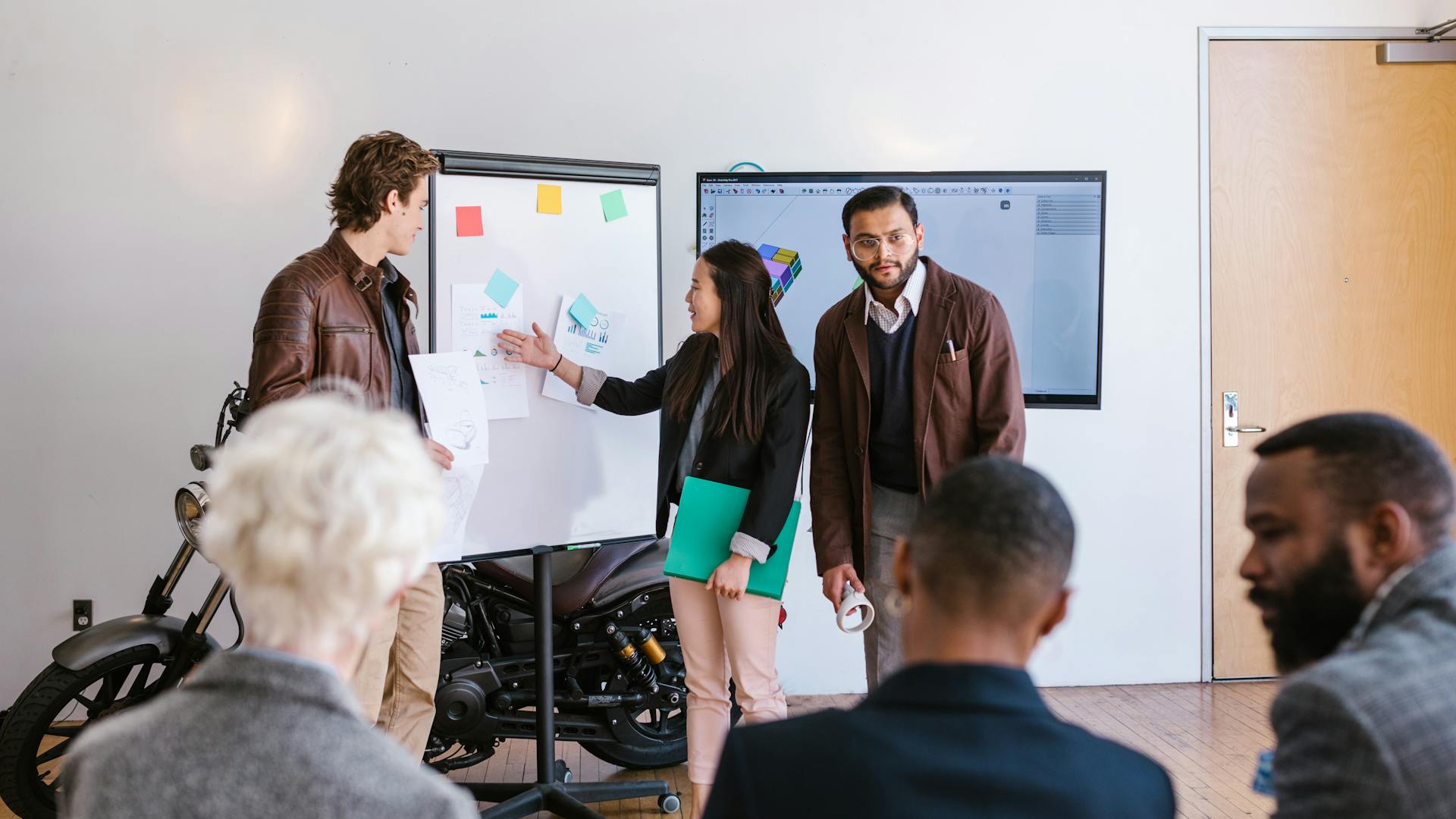 A multicultural team collaborates in an office, presenting ideas using a whiteboard and digital screen.