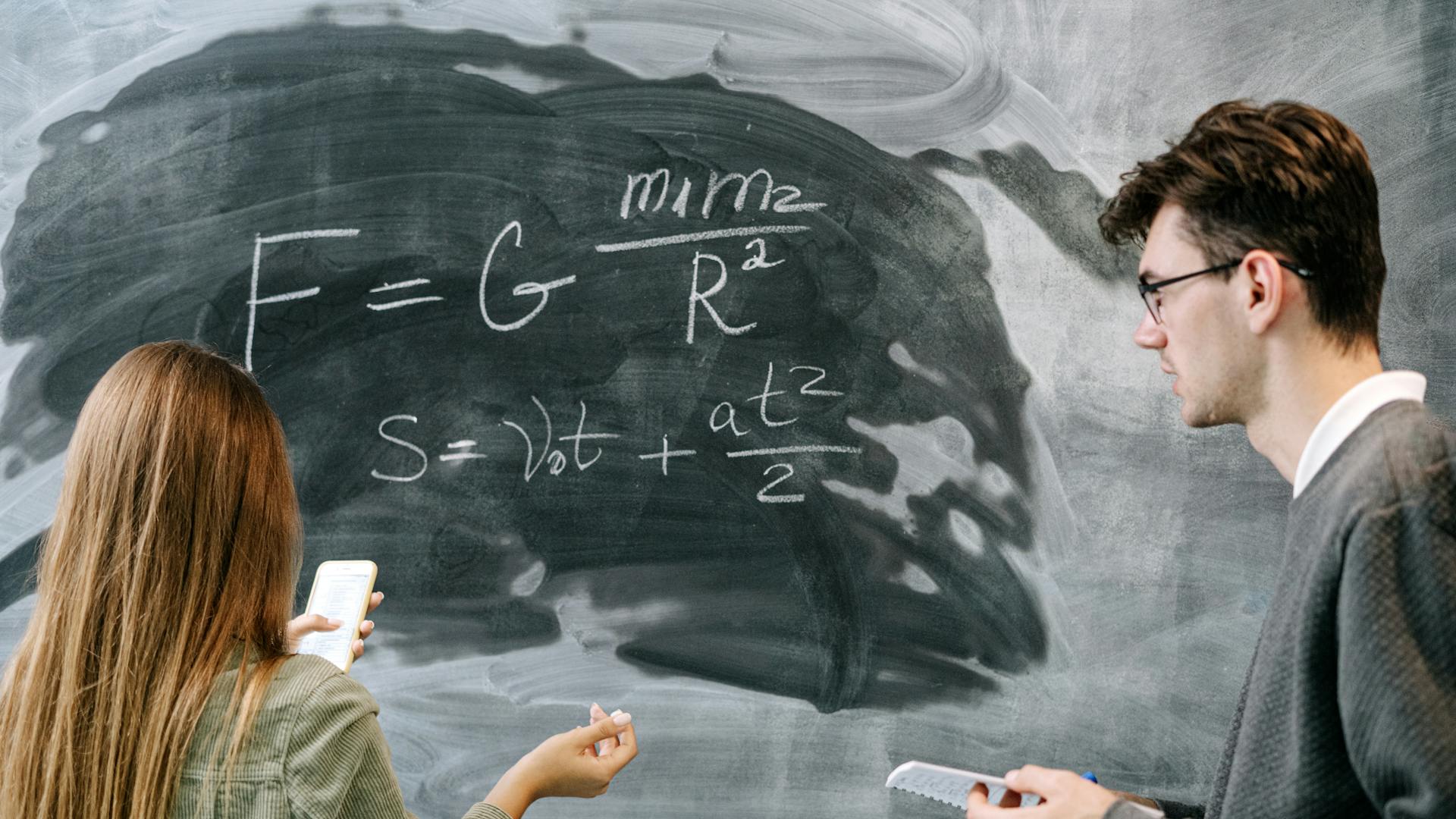 Two students discussing physics equations on a chalkboard in a classroom setting.