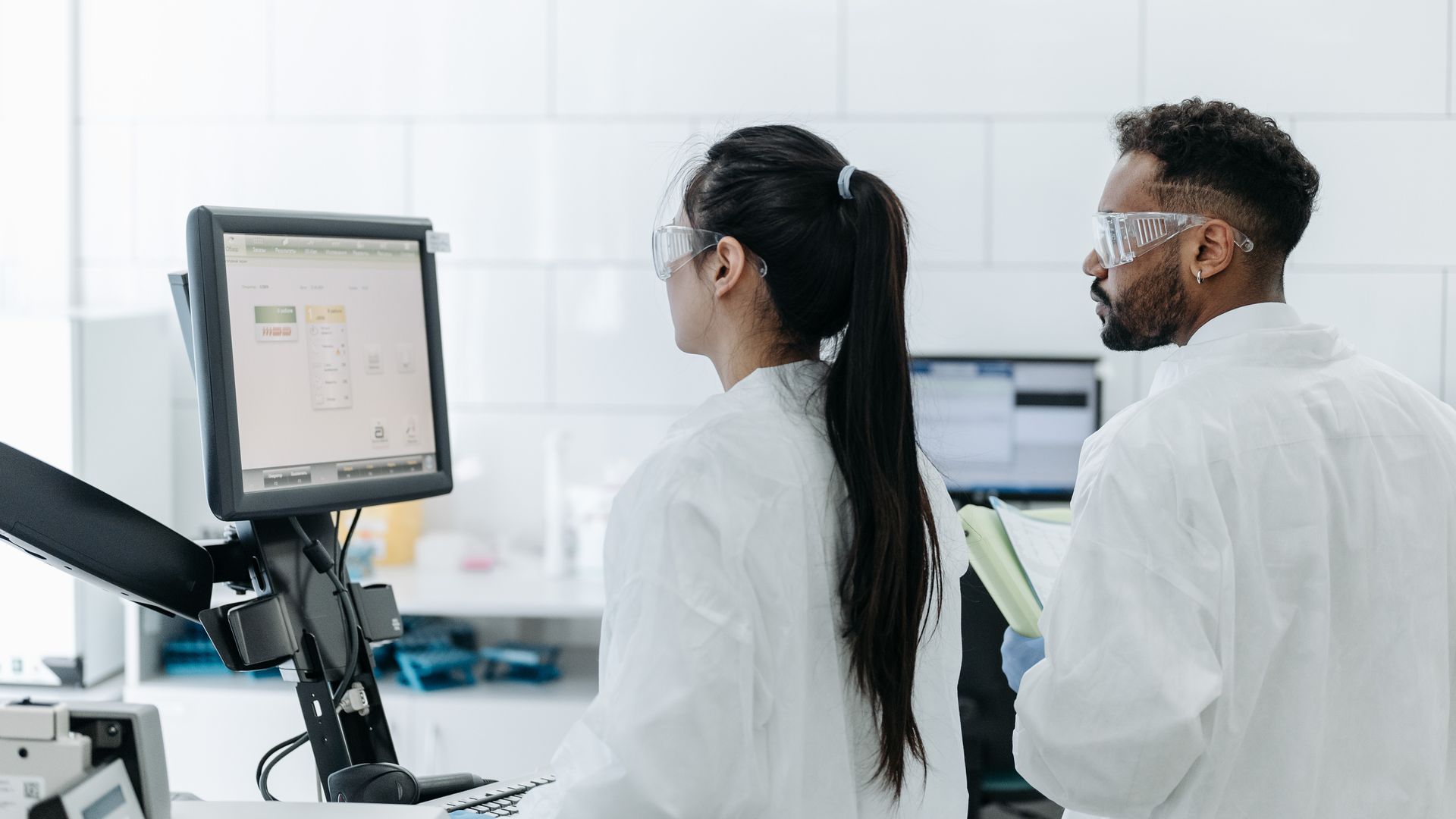 Two scientists wearing lab coats and goggles analyze data on a computer in a modern laboratory.